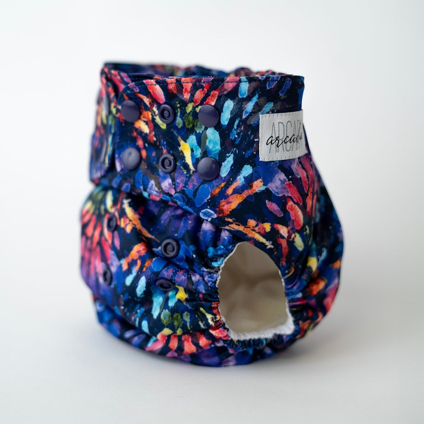 A pocket style cloth diaper at a slight angle, featuring a deep navy base with vivid splashes of blue, purple, pink, orange, and teal throughout. The abstract pattern feels painterly, with snap closures along the waist and rise for an adjustable fit. Soft leg elastics and a wide waistband are visible, along with the Arcadia’s Nursery logo label stitched at the hip.