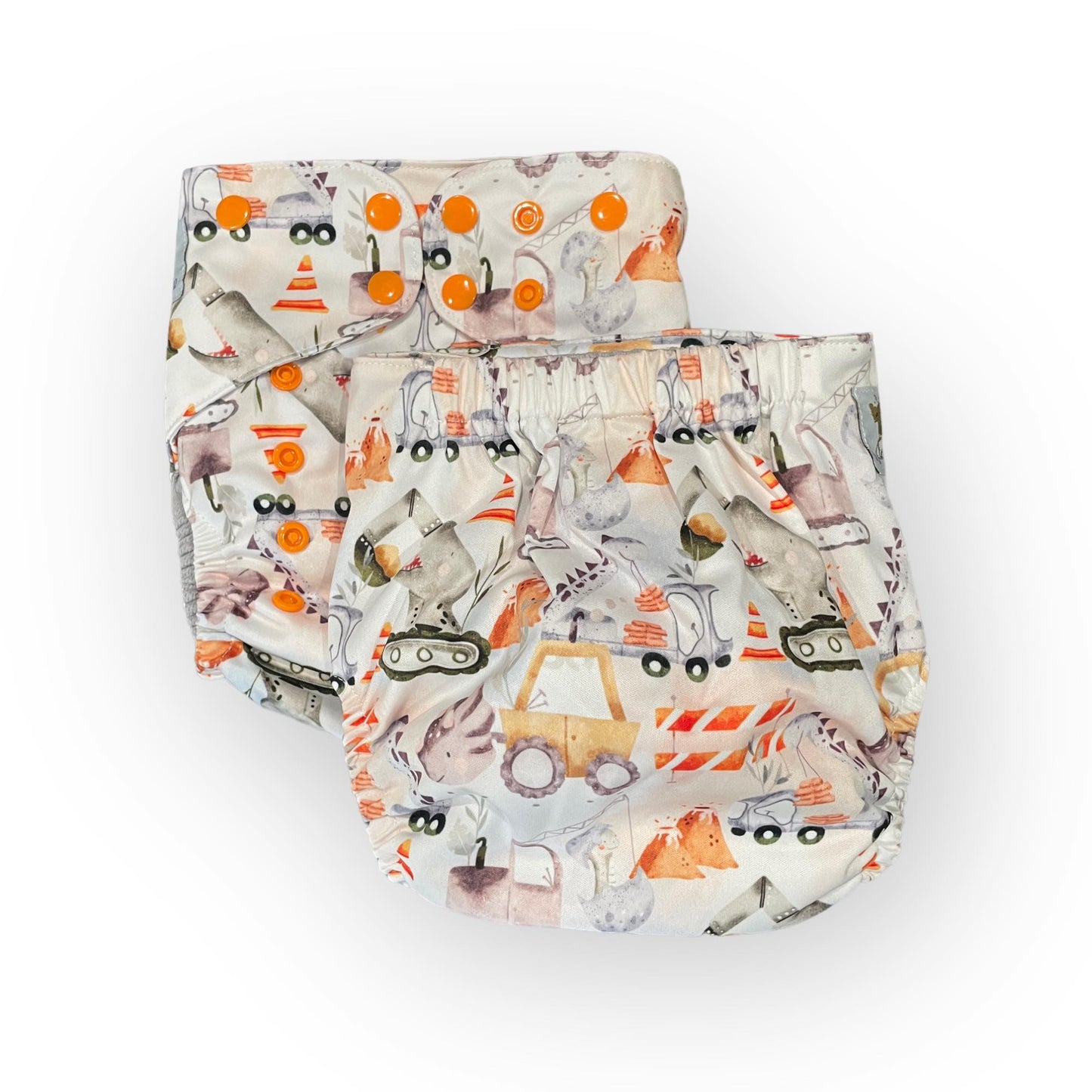 two pocket style wickham wild cloth diapers in the same print, one showing the front one showing the back. The print has a light background and features illustrations of construction vehicle-dinosaur hybrids, along with construction equipment like caution cones and road blocks.