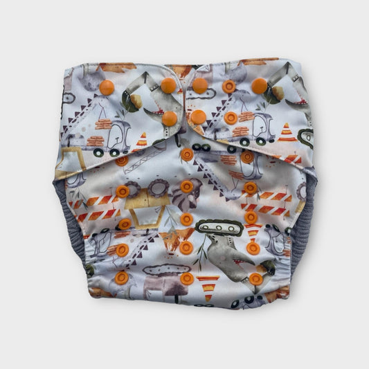 A wickham Wild pocket style cloth diaper. The print has a light background and features illustrations of construction vehicle-dinosaur hybrids, along with construction equipment like caution cones and road blocks. The snaps are orange.