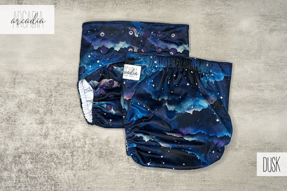 Two "Dusk" print pocket diapers, one showing the front and one the back, rest on a tabletop surface. The fabric features a nebula of navy, violet, & indigo with white stars... a white box in the top-left corner reads "arcadia" in script; a box in the bottom-right reads "DUSK" in tall sans-serif text. The diapers are finished with dark purple snaps & a white "arcadia" brand tag.