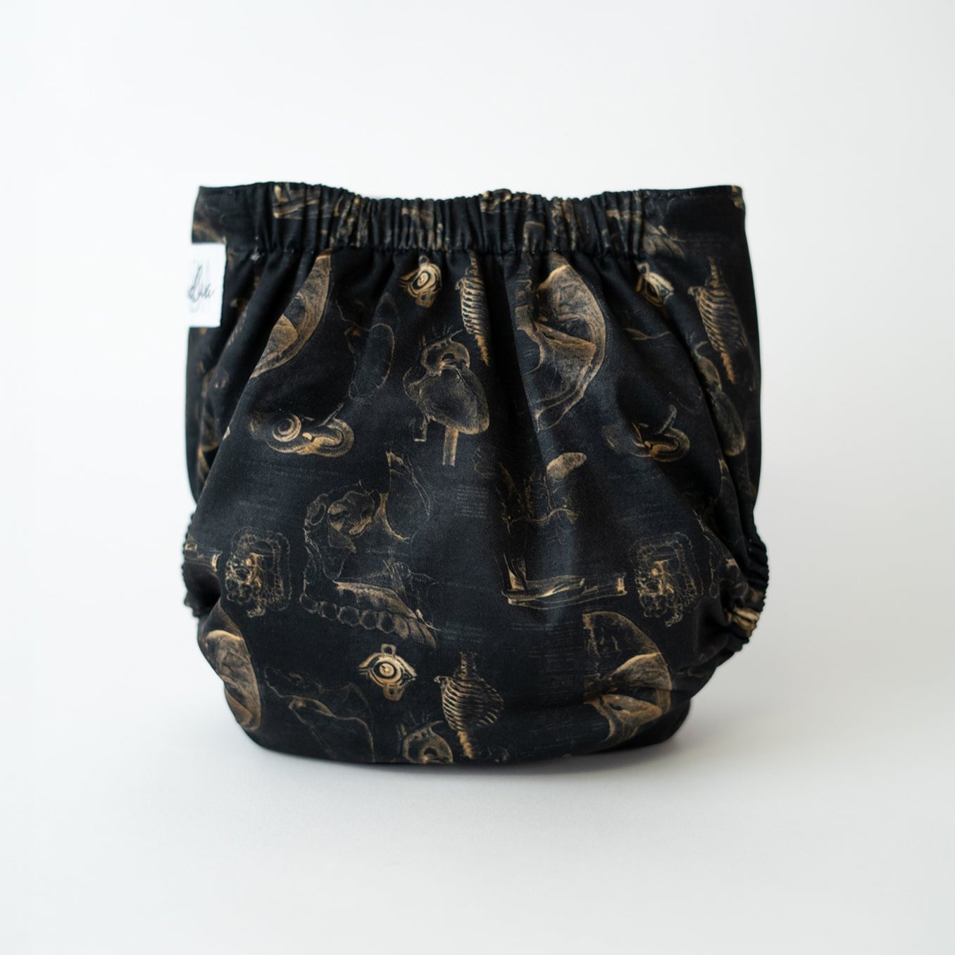 A pocket cloth diaper in the Fig 10 print shown from the back. The black fabric displays repeating sepia anatomical drawings of an eye, jaw, rib cage, and heart. The gathered elastic waistband is visible along the top, with the vintage textbook style artwork spread across the fabric.