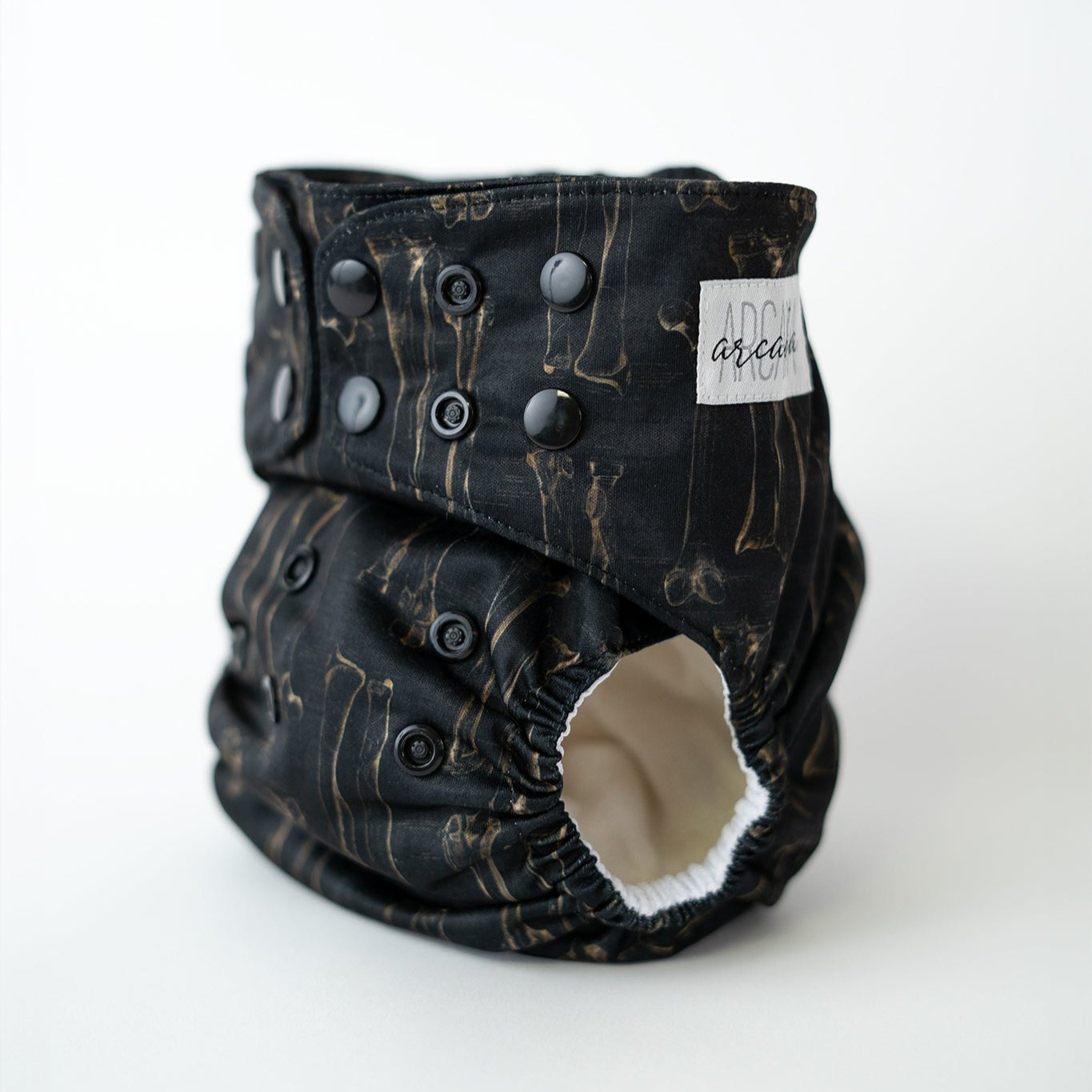 A reusable cloth diaper printed with spooky bones. The dark academia style depicts an illustration you may see in a vintage anatomy textbook. Black adjustable snaps are visible along the waist and rise. The leg opening reveals a white AWJ inner lining.