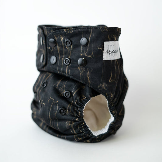 A reusable cloth diaper printed with spooky bones. The dark academia style depicts an illustration you may see in a vintage anatomy textbook. Black adjustable snaps are visible along the waist and rise. The leg opening reveals a white AWJ inner lining.