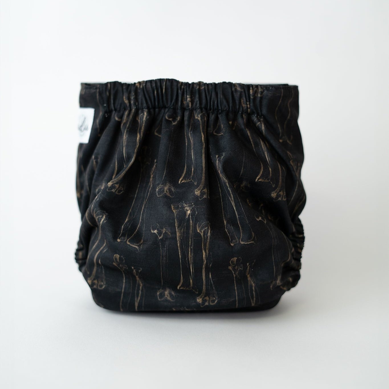 The back view of a pocket cloth diaper in the Fig 8 print, set against a white background. The black fabric features sepia anatomical illustrations of long bones arranged in a vintage textbook/ dark academia style. The gathered elastic waistband is visible along the top, with the bone illustrations repeating across the fabric.