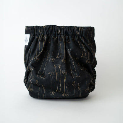 The back view of a pocket cloth diaper in the Fig 8 print, set against a white background. The black fabric features sepia anatomical illustrations of long bones arranged in a vintage textbook/ dark academia style. The gathered elastic waistband is visible along the top, with the bone illustrations repeating across the fabric.