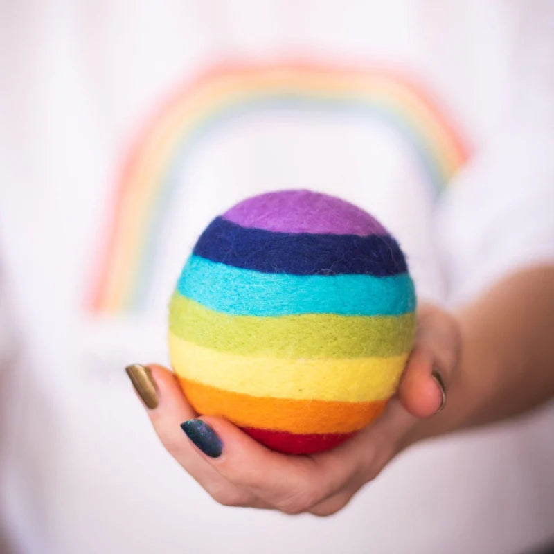 Colorful Pride striped wool dryer ball held by a hand with a blurred rainbow in the background
