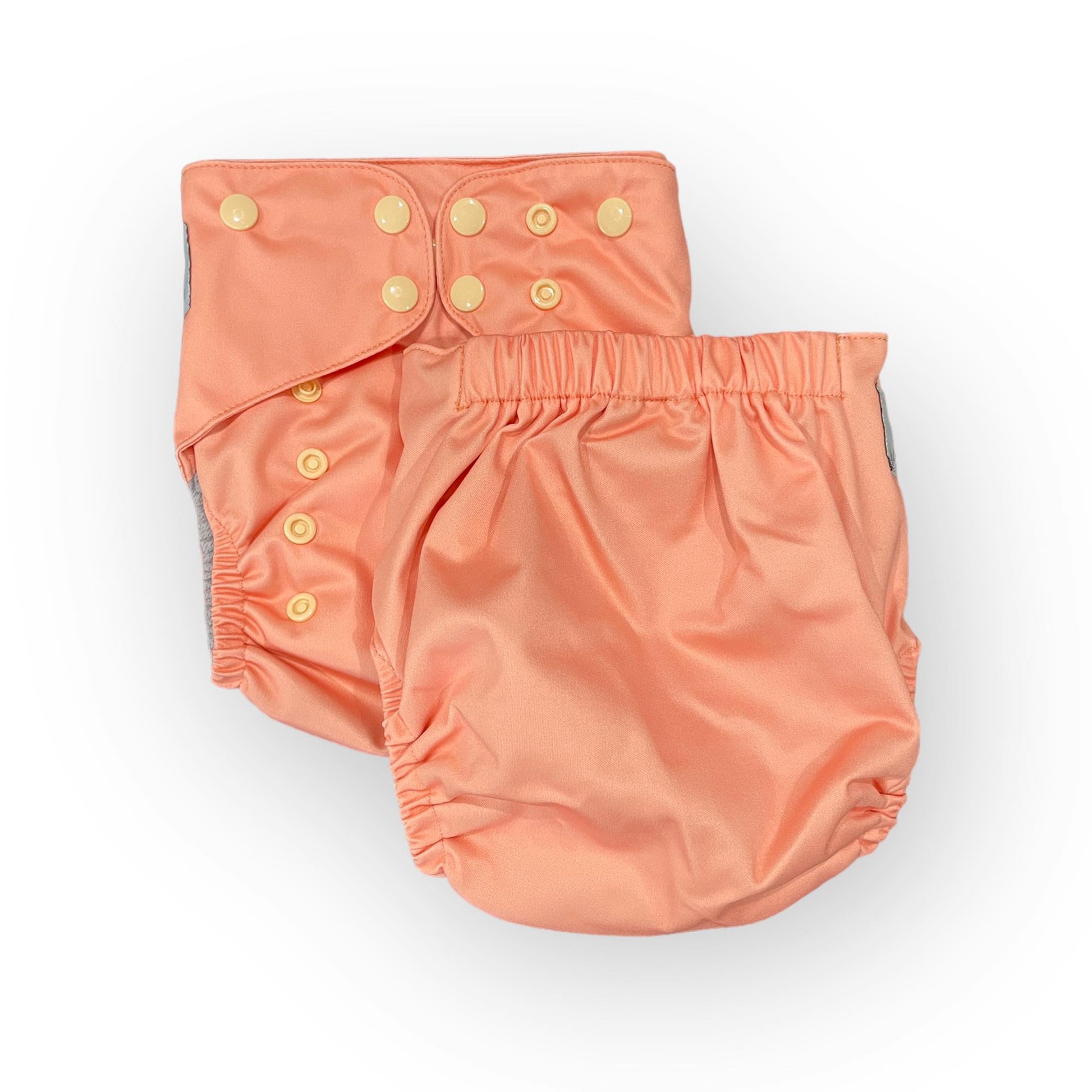 This image features two views of a peach-colored, reusable cloth diaper against a clean white background. On the left, the front of the diaper is shown, highlighting a grid of adjustable, cream-colored snaps used to customize the fit. The diaper on the right displays the back view, showcasing a thick, elasticized waistband designed for comfort and containment. Both views reveal the diaper's soft, slightly shimmering fabric and elastic leg openings