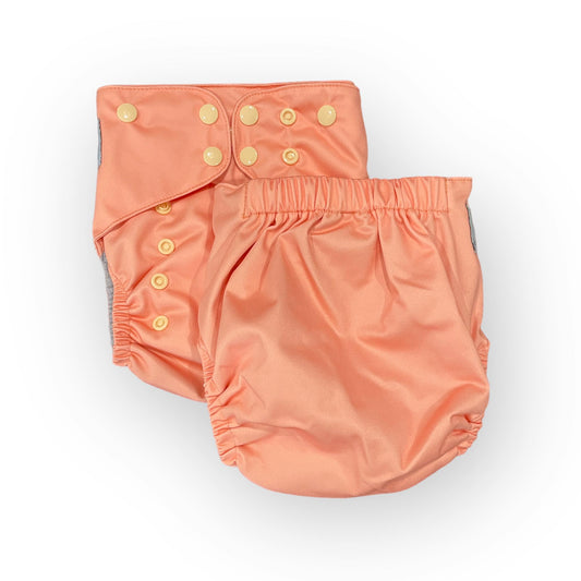 This image features two views of a peach-colored, reusable cloth diaper against a clean white background. On the left, the front of the diaper is shown, highlighting a grid of adjustable, cream-colored snaps used to customize the fit. The diaper on the right displays the back view, showcasing a thick, elasticized waistband designed for comfort and containment. Both views reveal the diaper's soft, slightly shimmering fabric and elastic leg openings
