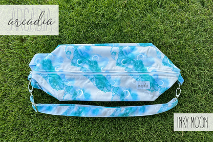 The "Inky Moon" patterned cloth diaper pod, with its blue alcohol ink moon design, is lying flat on vibrant green grass. The adjustable carrying strap is visible, along with silver hardware. The top zipper closure is partially visible. In the top left corner, the word "arcadia" is written in a script font. In the bottom right corner, the words "INKY MOON" are in a block font. The scene is illuminated by natural daylight.