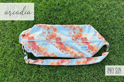 The "Inky Sun" patterned cloth diaper pod, with its orange, rust, and white abstract sun design, is lying flat on vibrant green grass. The adjustable carrying strap is visible, along with silver hardware. The top zipper closure is visible. In the top left corner, the word "arcadia" is written in a script font. In the bottom right corner, the words "INKY SUN" are in a block font. The scene is illuminated by natural daylight.