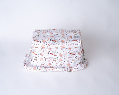 A photo of a cloth diaper pod against a plain white backrop. The pod has a white background, with iillustrations of UFOs, rockets, stars, rainbows, and moons in shades of rust and gold. The detatchable shoulder strap is visible with it's adjustable slider and metal hardware, along with the 2 zipper compartments. There is a white arcadia's logo tag stitched onto the fabric on the right side of the top of the bag.
