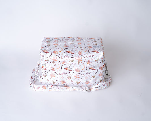 A photo of a cloth diaper pod against a plain white backrop. The pod has a white background, with iillustrations of UFOs, rockets, stars, rainbows, and moons in shades of rust and gold. The detatchable shoulder strap is visible with it's adjustable slider and metal hardware, along with the 2 zipper compartments. There is a white arcadia's logo tag stitched onto the fabric on the right side of the top of the bag.
