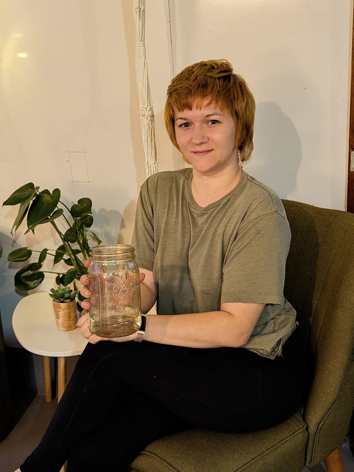 Kristin, owner of Arcadia's Nursery LLC, sitting on a chair holding a glass of water, with a plant in the background.