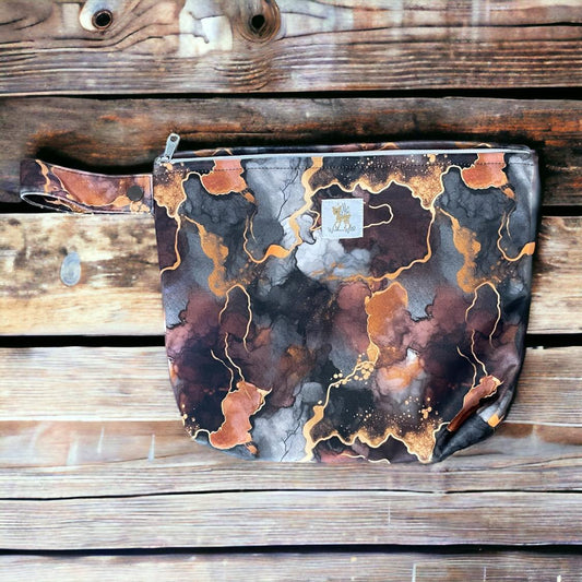 a small travel pouch in a dark and moody watercolor/alcohol ink style print featuring dark grey, burgundy, orange and gold. you can see the wickham wild logo tag stitched onto the front. there is a wrist strap with snap closure and ykk zipper closure for the pouch. the pouch is laid on a wooden background.