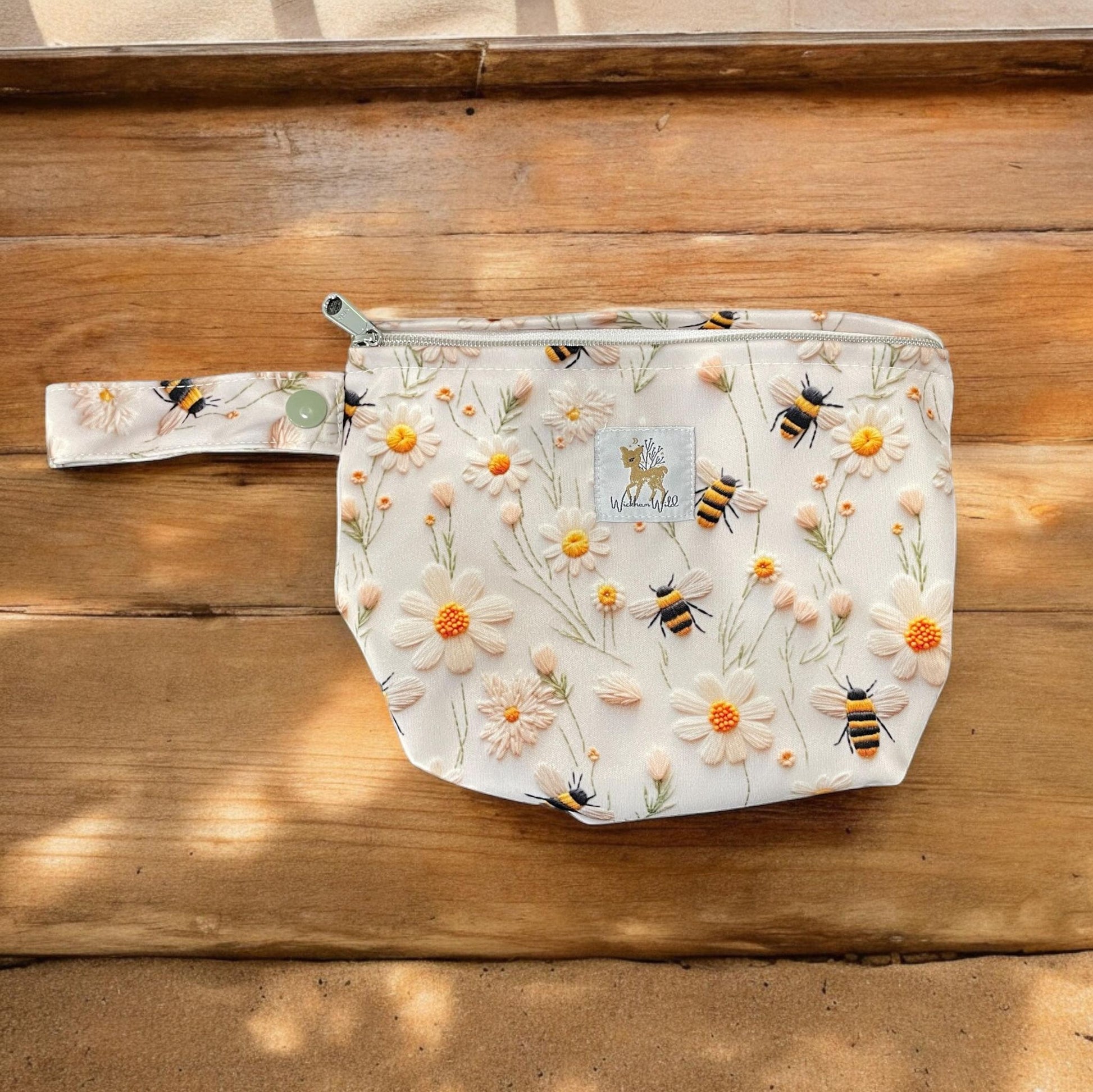 a small andiamo pouch in a bee and flower print. the background of the print is light cream with faux embridery style bees and white flowers, laying on a wood background.