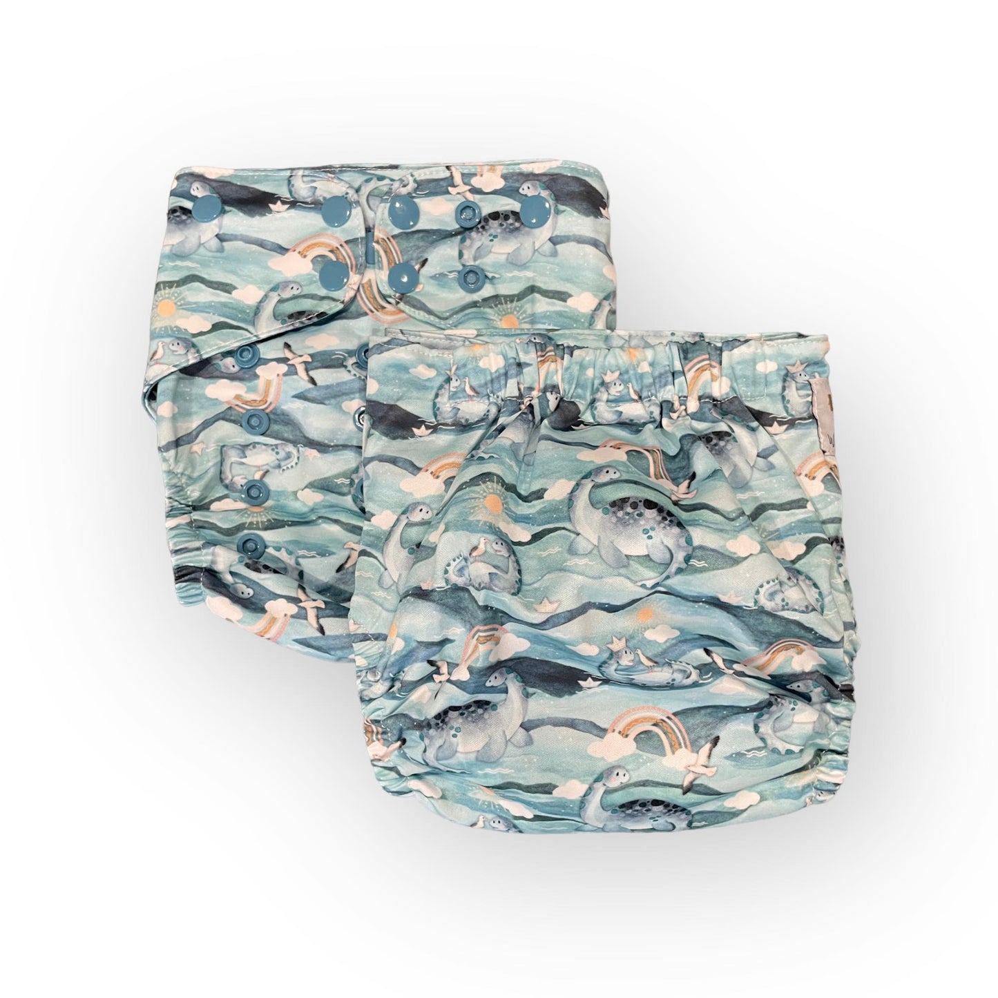 Photo showing 2 pocket cloth diapers with the same print, one from the front and one from the back. They are blue pocket cloth diapers with illustrations of sea gulls, plesiosaurus, brown rainbows, clouds, the sun and water.