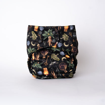 The front view of an Arcadia’s Nursery pocket cloth diaper in the Oddities print, displayed against a white background. The artwork highlights mandrake illustrations alongside potion bottles, botanicals, and ritual objects associated with green witchcraft. Black snaps provide adjustable sizing across the waist and rise, while the black AWJ interior and black Arcadia’s logo tag reinforce the cohesive, dark aesthetic.