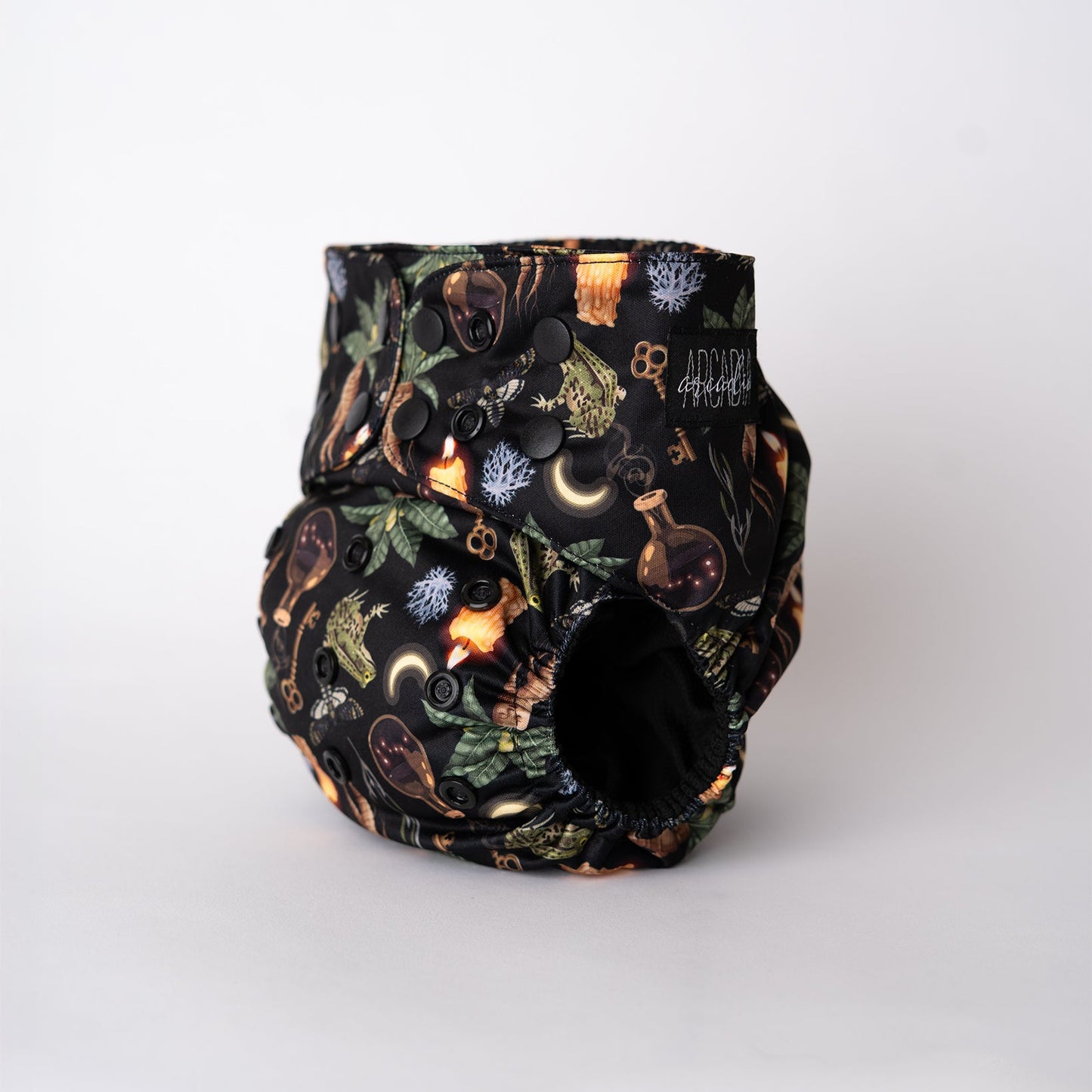 A pocket style cloth diaper from Arcadia’s Nursery shown at a slight front angle in the Oddities print. The dark, richly detailed pattern features illustrations tied to green witchcraft, including mandrake roots, apothecary, spell jars, candles, keys, and mystical tools. Black snap closures line the waist and rise, the black athletic wicking jersey interior is visible at the leg opening, and a black Arcadia’s Nursery logo tag is stitched at the hip. Soft elastics create a contoured fit.