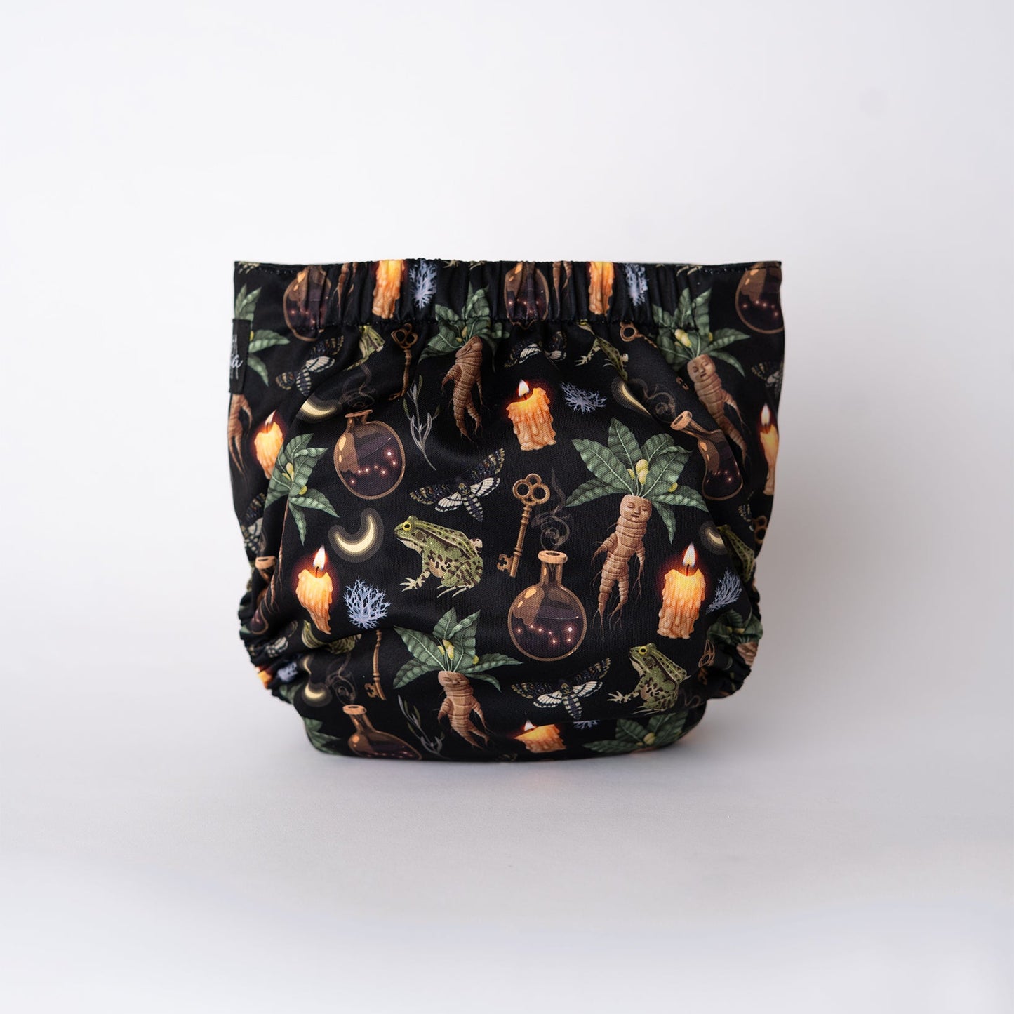The back view of a pocket style cloth diaper in the Oddities print from Arcadia’s Nursery. Mandrake roots, herbs, candles, and apothecary imagery repeat across the full seat on a deep black base. Gathered elastic at the waist and legs creates a snug, comfortable shape, with the uninterrupted print emphasizing the moody, witchy theme.