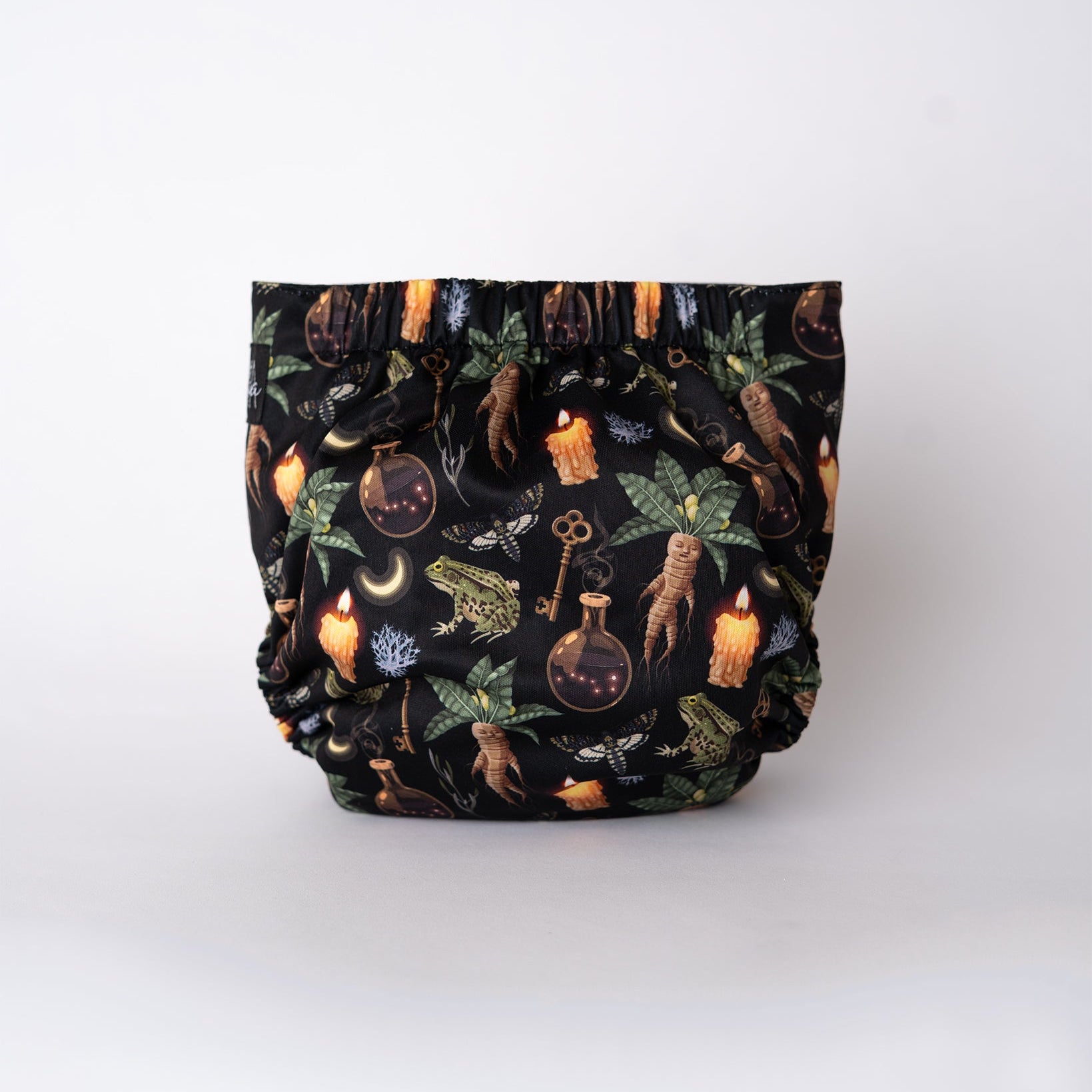 The back view of a pocket style cloth diaper in the Oddities print from Arcadia’s Nursery. Mandrake roots, herbs, candles, and apothecary imagery repeat across the full seat on a deep black base. Gathered elastic at the waist and legs creates a snug, comfortable shape, with the uninterrupted print emphasizing the moody, witchy theme.