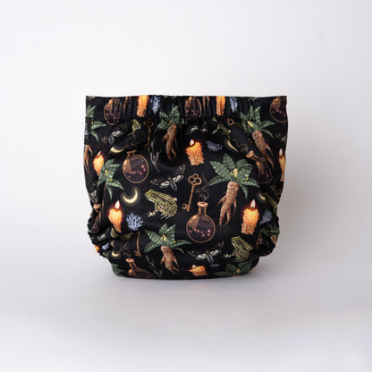 The back view of a pocket style cloth diaper in the Oddities print from Arcadia’s Nursery. Mandrake roots, herbs, candles, and apothecary imagery repeat across the full seat on a deep black base. Gathered elastic at the waist and legs creates a snug, comfortable shape, with the uninterrupted print emphasizing the moody, witchy theme.