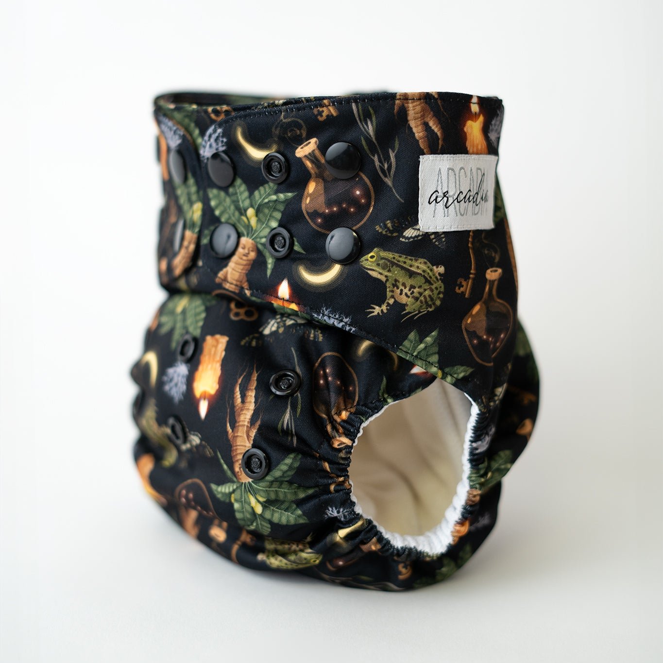 A photo of a reusable cloth diaper featuring a witchcraft inspired print. It features illustrations of spell jars, frogs, moths, mandrakes, keys, and moons. This whimsical, botanical, green witch inspired pattern is perfect for spooky spiritual moms. front angle view.
