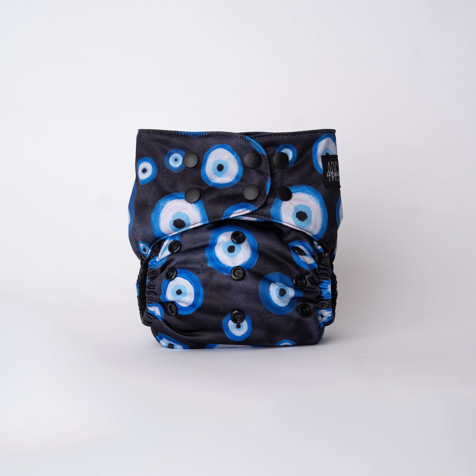 A photo of a cloth diaper standing up with a plain white background. The pattern on the cloth diaper is a light black watercolor, with hand painted Eye illustrations. The eyes are blue, black, and white. This symbol is commonly reffered to as "evil eye" but is known as Mati or Nazar. The snaps are matte black and the Arcadia's Nursery Tag is black. Front view.