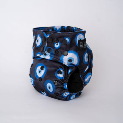 A photo of a cloth diaper standing up with a plain white background. The pattern on the cloth diaper is a light black watercolor, with hand painted Eye illustrations. The eyes are blue, black, and white. This symbol is commonly reffered to as "evil eye" but is known as Mati or Nazar. The snaps are matte black and the Arcadia's Nursery Tag is black. Front angle view of the diaper.