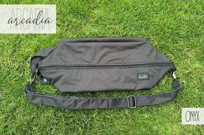 A black cloth diaper pod shown flat, outdoors on the grass. The adjustable shoulder strap with metal hardware are visible, along with the carrying handle with snaps, and black zipper closure.  There is a black arcadia's nursery logo tag sewn onto the right side of the pod. The entire photo is light by natural daylight. In the upper lefthand corner is the arcadias nursery logo, and in the bottom right hand of the graphic there is "ONYX" in a sans seriff font.