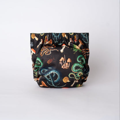 The back of a pocket style cloth diaper is displayed against a white backdrop, showing gathered elastic along the waistband and leg openings. The print continues uninterrupted across the back panel, featuring serpents and forest-inspired details in green, gold, and rust tones set against black. Illustrations of beetles, mushrooms, snakes, and preying mantis are visible. 