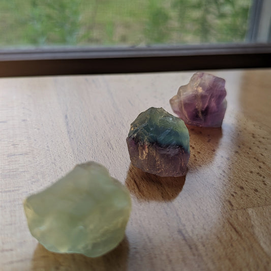 Three raw crystal stones with blue, purple, green, and clear colorations on a wooden surface.