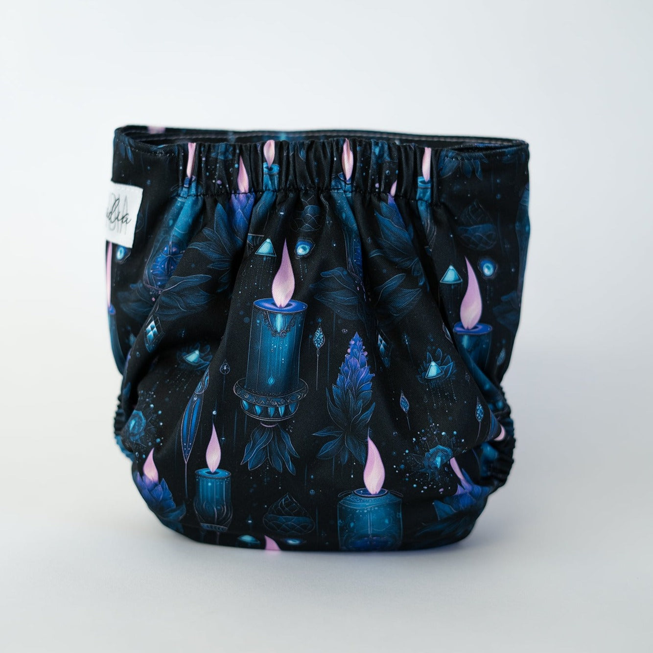The back view of a pocket style cloth diaper in the Séance print, photographed against a clean white background. The background of the fabric is a deep blackish blue, with illustrations of lit blue candles with a pink flame.