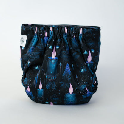 The back view of a pocket style cloth diaper in the Séance print, photographed against a clean white background. The background of the fabric is a deep blackish blue, with illustrations of lit blue candles with a pink flame.