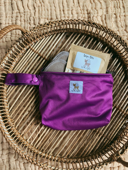 A photo of a purple small andiamo pouch, with the zipper open demonstrating what contents can fit. this image shows a packet of cloth wipe bits and cloth wipes.