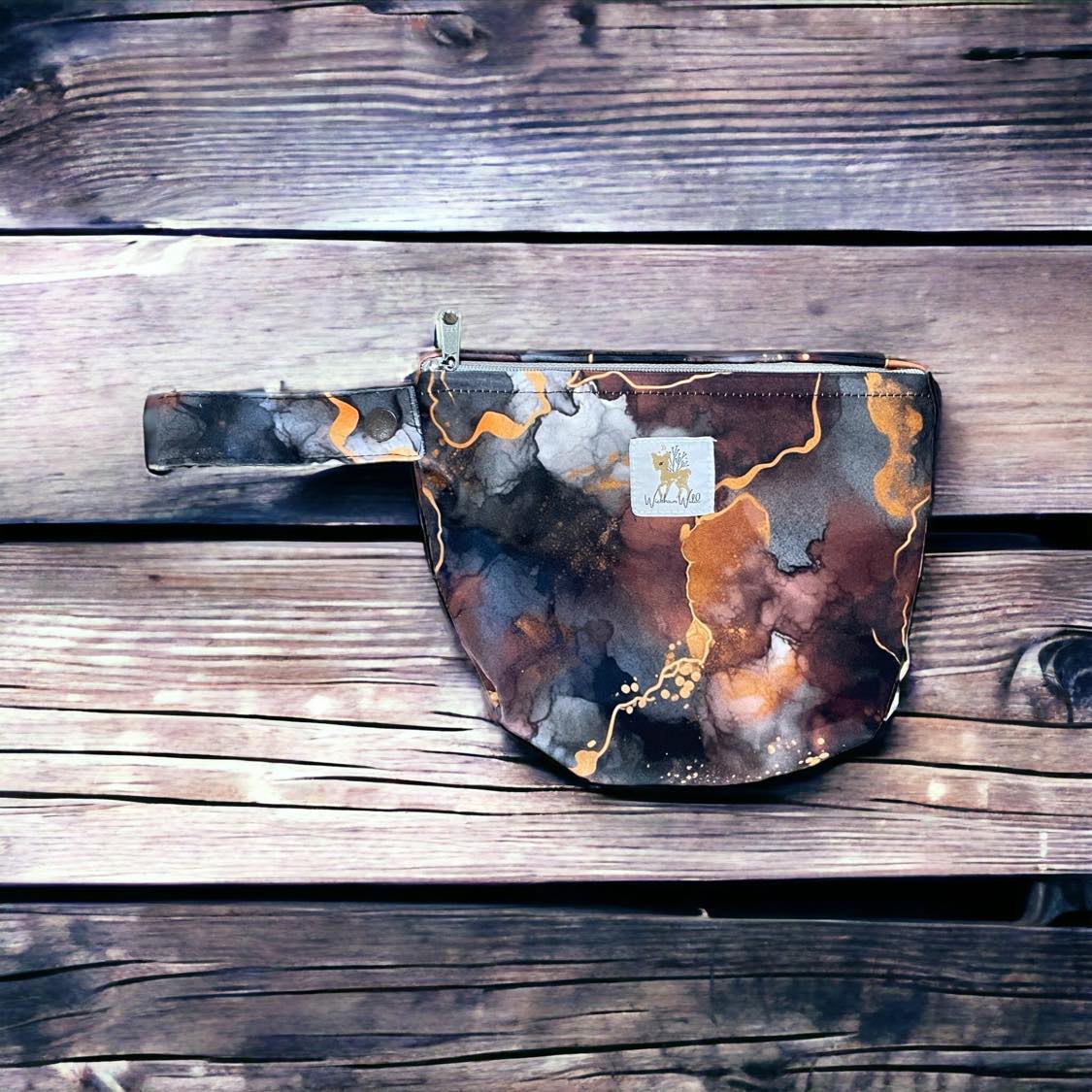 a mini travel pouch in a dark and moody watercolor/alcohol ink style print featuring dark grey, burgundy, orange and gold. you can see the wickham wild logo tag stitched onto the front. there is a wrist strap with snap closure and ykk zipper closure for the top of pouch. the pouch is laid on a wooden background.