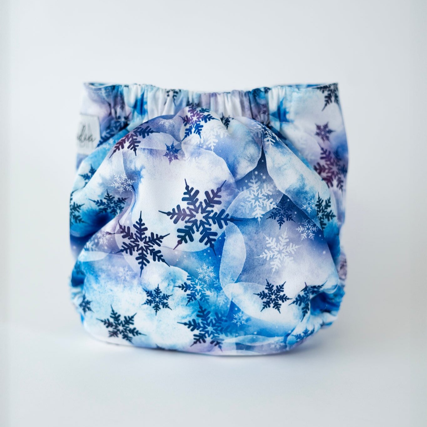 The back view of an Arcadia’s Nursery pocket style cloth diaper shown against a clean white background. The print features layered watercolor style clouds in soft shades of icy blue, slate, and white, overlaid with delicate snowflake motifs in navy and indigo. Gathered elastic along the waist and legs creates a gently contoured shape.