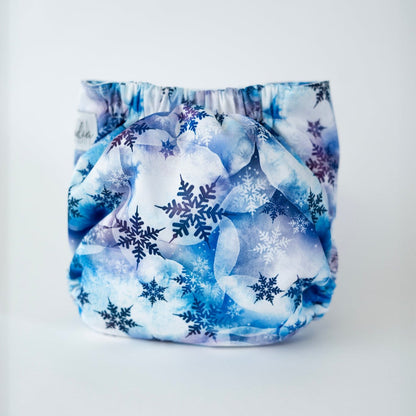 The back view of an Arcadia’s Nursery pocket style cloth diaper shown against a clean white background. The print features layered watercolor style clouds in soft shades of icy blue, slate, and white, overlaid with delicate snowflake motifs in navy and indigo. Gathered elastic along the waist and legs creates a gently contoured shape.