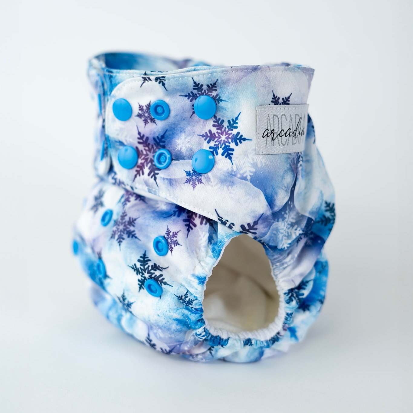 An angled front view of an Arcadia’s Nursery pocket style cloth diaper featuring a cool toned winter print. Blue snap closures line the waist and rise for an adjustable fit, complementing the watercolor white and blue cloud background and scattered blue and indigo snowflake designs. The curved leg opening with soft elastic and the Arcadia’s Nursery woven label are visible, highlighting both the functional construction and the crisp, seasonal aesthetic.