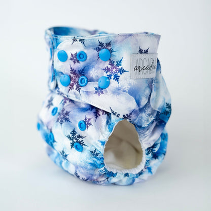 An angled front view of an Arcadia’s Nursery pocket style cloth diaper featuring a cool toned winter print. Blue snap closures line the waist and rise for an adjustable fit, complementing the watercolor white and blue cloud background and scattered blue and indigo snowflake designs. The curved leg opening with soft elastic and the Arcadia’s Nursery woven label are visible, highlighting both the functional construction and the crisp, seasonal aesthetic.