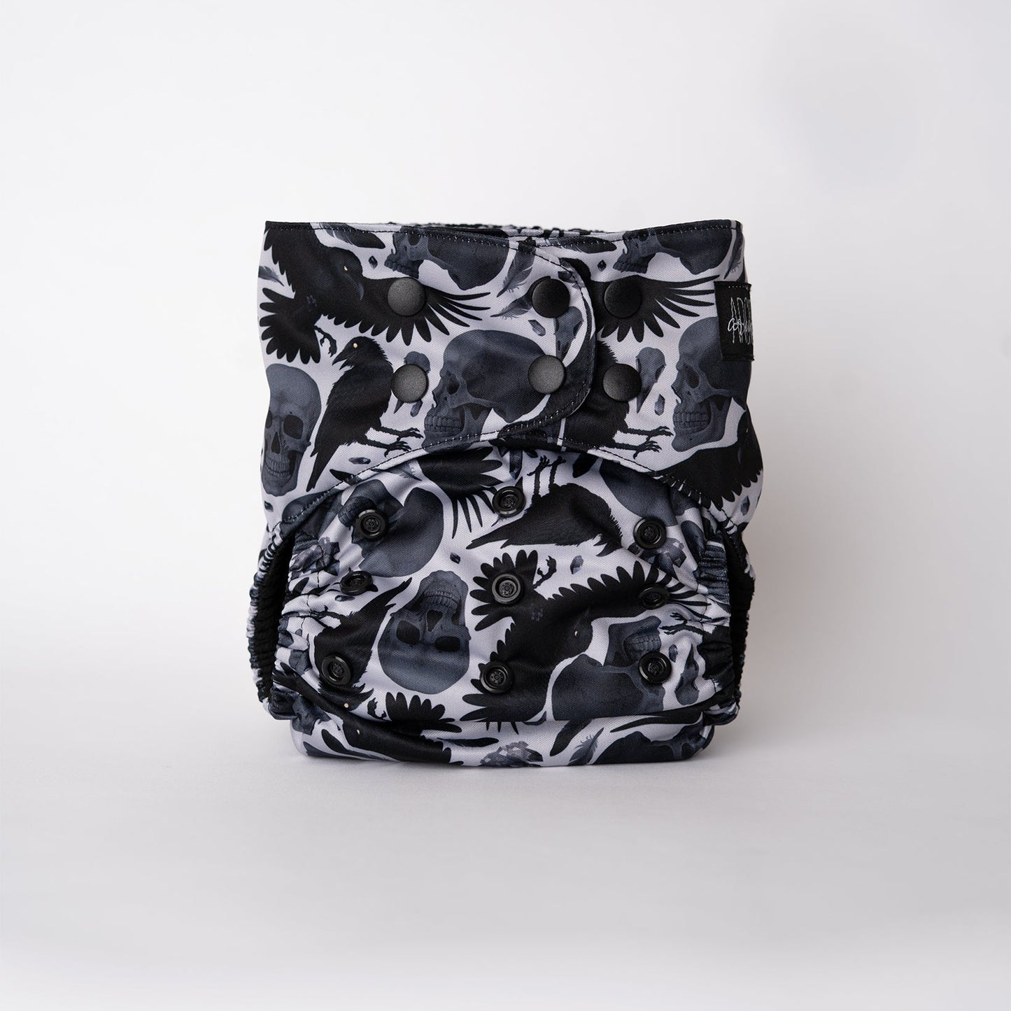 The front view of an Arcadia’s Nursery pocket cloth diaper displayed against a white background. The Raven print showcases black ravens, skull motifs, and scattered feathers arranged across the front panel. Snap closures along the waist and rise allow for a customizable fit, while the dramatic, witchy artwork remains the focal point.