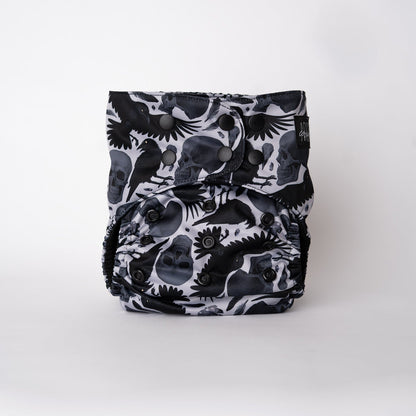 The front view of an Arcadia’s Nursery pocket cloth diaper displayed against a white background. The Raven print showcases black ravens, skull motifs, and scattered feathers arranged across the front panel. Snap closures along the waist and rise allow for a customizable fit, while the dramatic, witchy artwork remains the focal point.