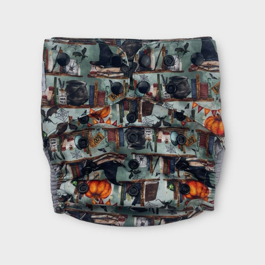 a reusable pocket style diaper with a green background depicting bookshelves filled with spooky motifs. There are books on the shelves along with witches and cauldrons, a spider in a jar, pumpkin, dragon, spider webs, and spell jars. the snaps are black.