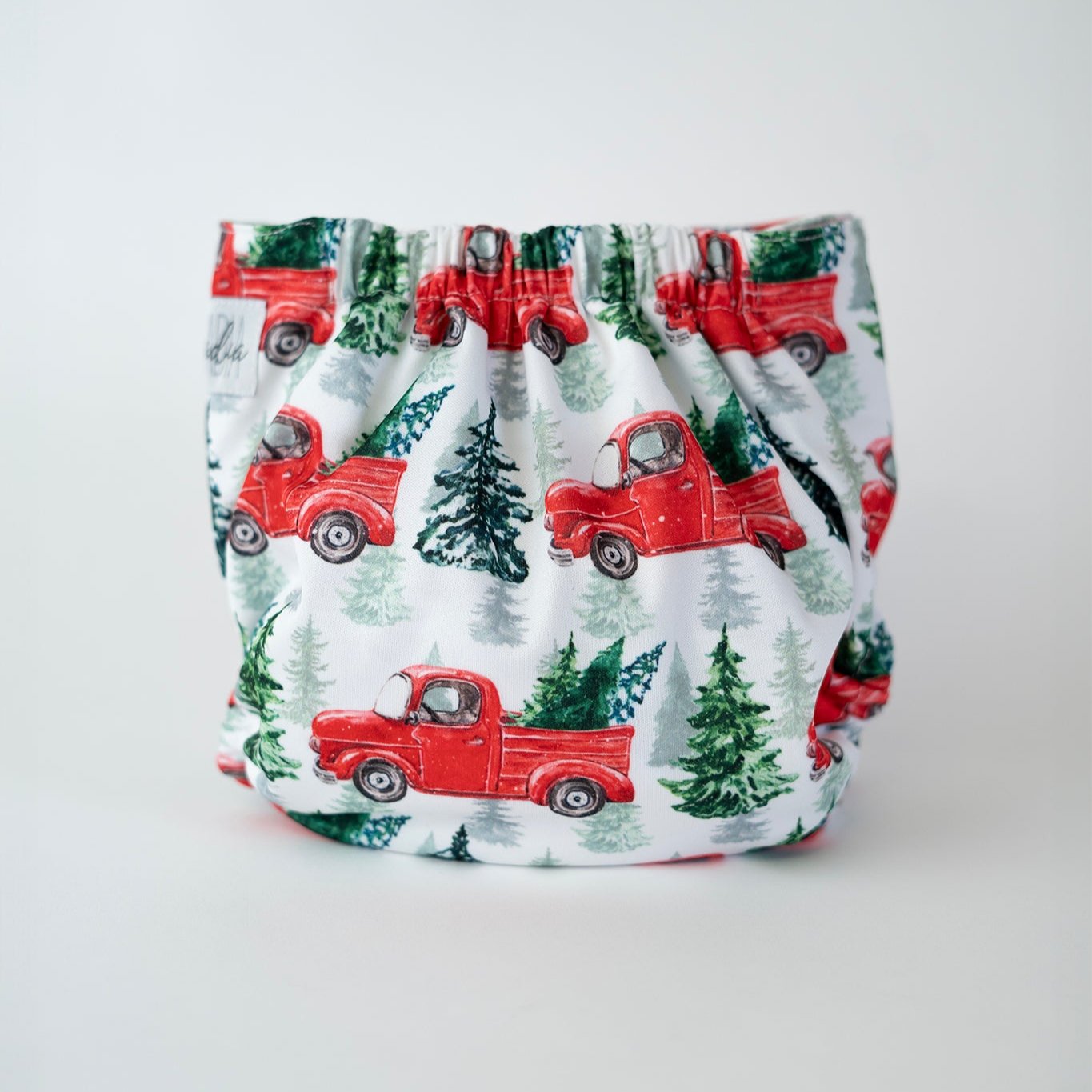 The back view of an Arcadia’s Nursery pocket style cloth diaper shown against a clean white background. The print features a white background, and illustrations of vintage red trucks with trees in the truck bed. Gathered elastic along the waist and legs creates a gently contoured shape, while the fabric displays the winter inspired pattern uninterrupted.