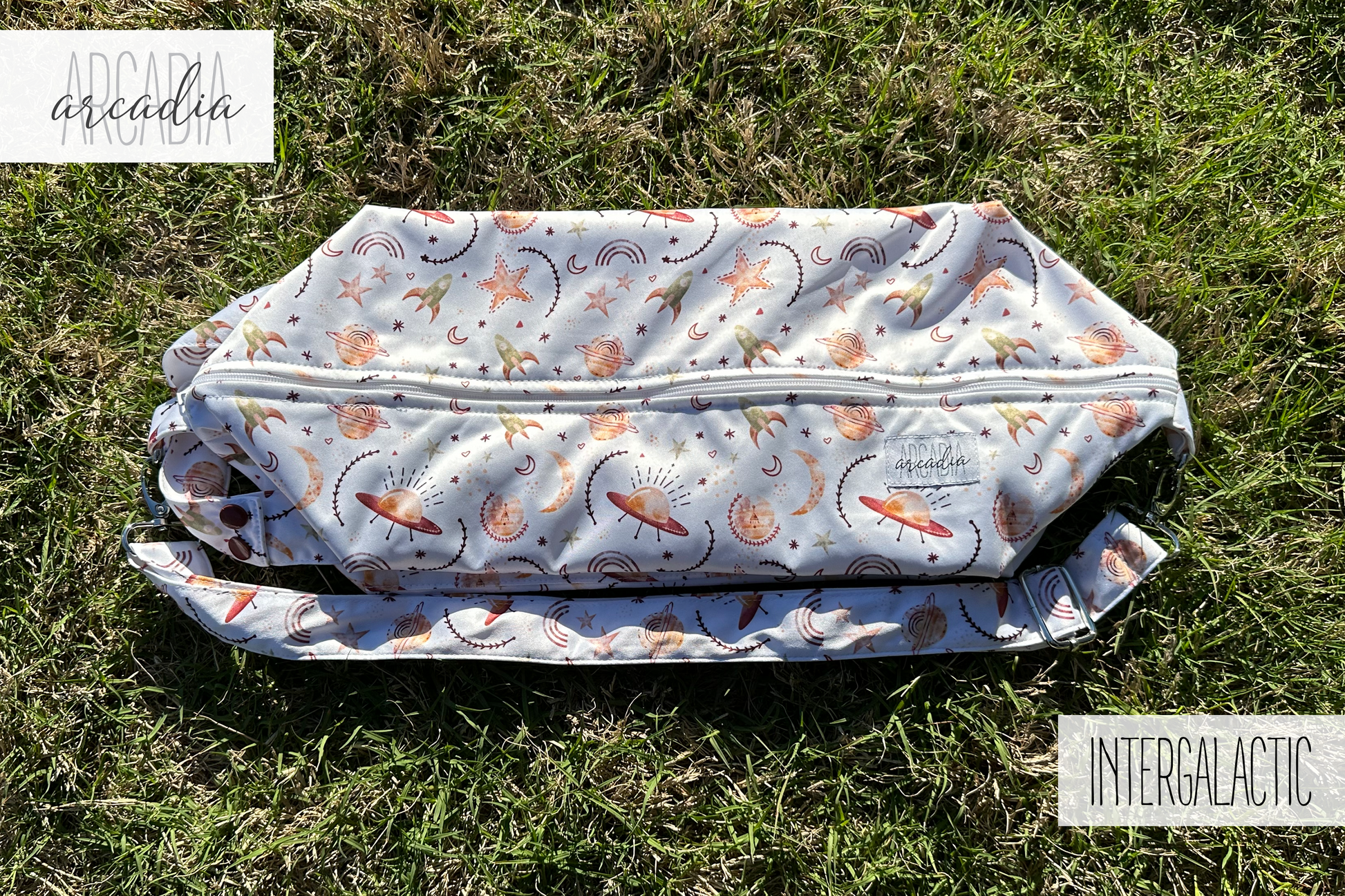 A photo of a cloth diaper pod laying flat on grass. The pod has a white background, with watercolor imagery of UFOs, rockets, stars, rainbows, and moons in rusty and golden hues. You can see one of the zipper compartments and the adjustable shoulder strap along with the carrying handle. In the upper left corner is an arcadia's logo, and in the lower right text that reads "INTERGALACTIC". The photo is illuminated by natural daylight.