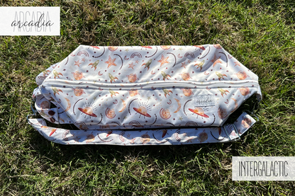A photo of a cloth diaper pod laying flat on grass. The pod has a white background, with watercolor imagery of UFOs, rockets, stars, rainbows, and moons in rusty and golden hues. You can see one of the zipper compartments and the adjustable shoulder strap along with the carrying handle. In the upper left corner is an arcadia's logo, and in the lower right text that reads "INTERGALACTIC". The photo is illuminated by natural daylight.